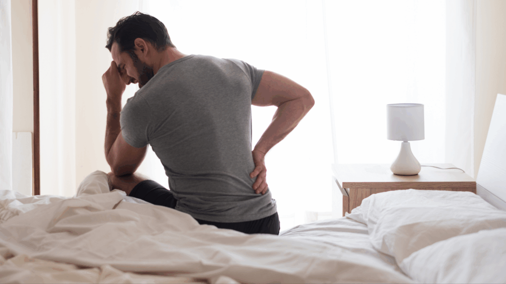 Man sitting on bed in pain, holding his lower back with one hand and his forehead with the other.