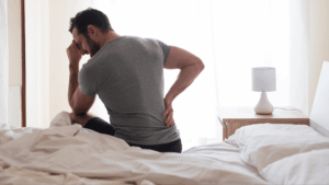 Man sitting on bed in pain, holding his lower back with one hand and his forehead with the other.