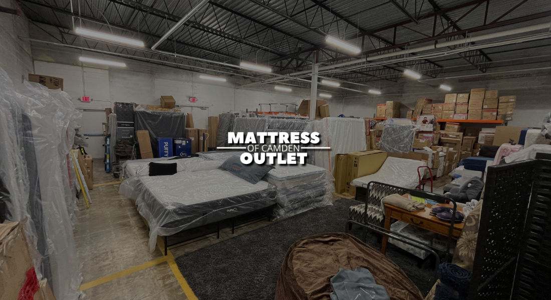 Mattresses and bedding in a warehouse labeled Mattress of Camden Outlet with boxes and furniture.