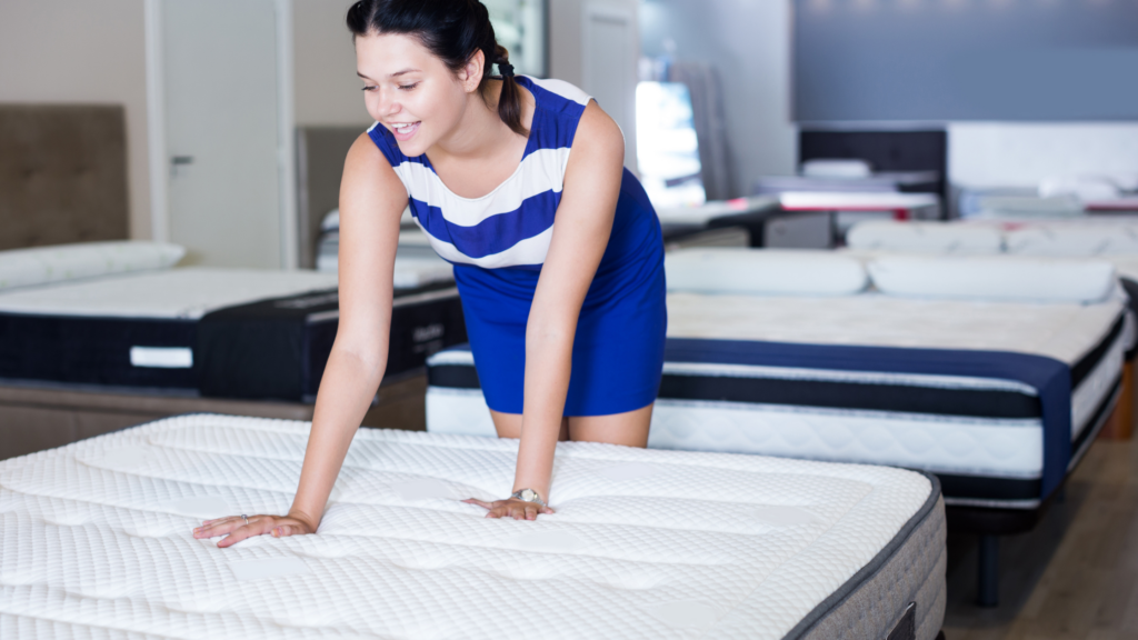 Woman in a blue and white dress presses on a mattress in a showroom, testing its firmness.
