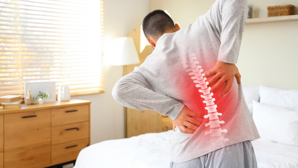 Man holding his lower back in pain with a highlighted spine illustration, standing beside a bed in a bright room.