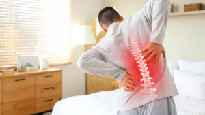 Man holding his lower back in pain with a highlighted spine illustration, standing beside a bed in a bright room.