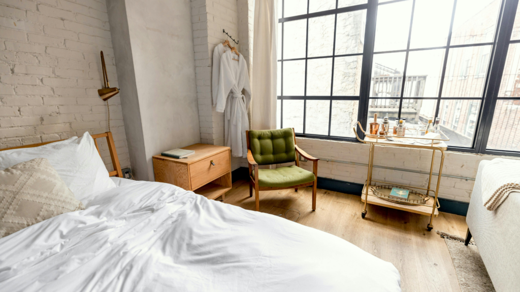 Bright bedroom with an unmade bed, green chair, bathrobes, and a drink cart by large windows.